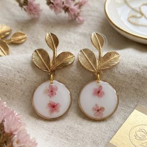Boucles d'oreilles dorees avec motif de feuilles et pendentif rond contentant de veritables myosotis roses figes dans la resine sur fond blanc, posees sur un tissu en lin a cote de fleurs sechees.