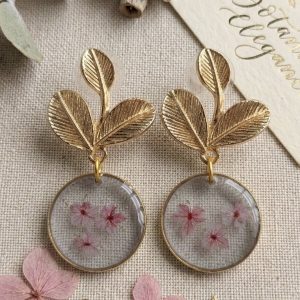 Boucles d'oreilles dorees en forme de feuilles avec des pendentifs ronds transparents laissant apparaitre trois myosotis roses, presentees sur un fond en lin avec des fleurs sechees et une etiquette.