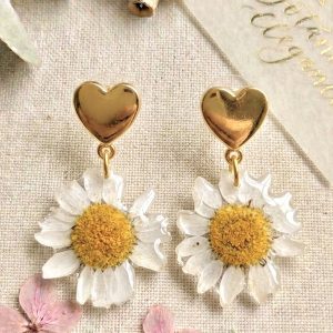 Boucles d'oreilles dorees en forme de coeurs avec de veritables marguerites suspendues, figees dans une resine transparente, posees sur un tissu en lin avec des fleurs roses