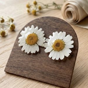 Paire de boucles d'oreilles artisanales en forme de marguerites reelles figees dans la resine, presentees sur un support en bois fonce avec des fleurs de camomille sechees en arriere-plan