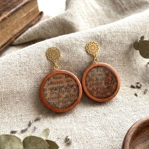 Paire de boucles d'oreilles rondes en bois naturel contenant des fragments de textes anciens sous resine, surmontees s'une puce d'oreille doree en forme de rosace ciselee, posees sur un lin beige a cote d'un vieux livre.