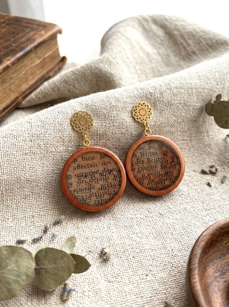 Paire de boucles d'oreilles rondes en bois naturel contenant des fragments de textes anciens sous resine, surmontees s'une puce d'oreille doree en forme de rosace ciselee, posees sur un lin beige a cote d'un vieux livre.