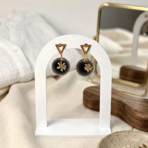 Boucles d'oreilles artisanales avec de véritables fleurs de gliksja blanches figées dans une résine noire, suspendues sur un présentoir blanc en forme d'arche devant une chemise en lin clair.