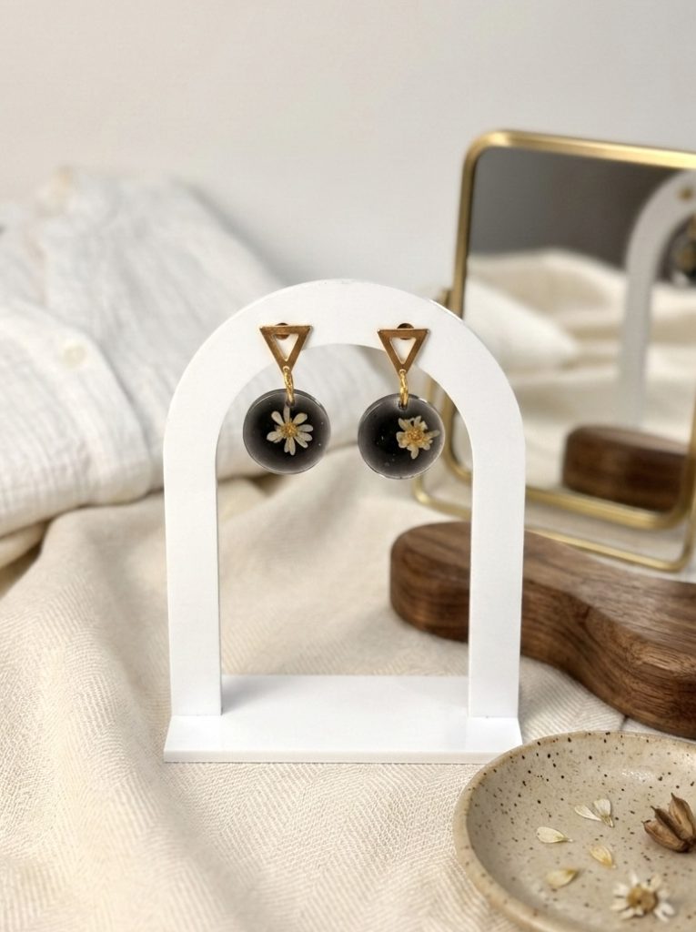 Boucles d'oreilles artisanales avec de véritables fleurs de gliksja blanches figées dans une résine noire, suspendues sur un présentoir blanc en forme d'arche devant une chemise en lin clair.