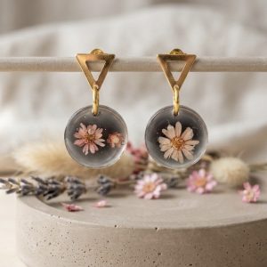 Gros plan sur des boucles d'oreilles artisanales avec des fleurs de gliksja roses dans une résine noire, posées sur un tissu en lin clair à côté d'un bol en céramique et d'éléments en bois.