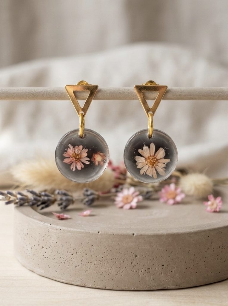 Gros plan sur des boucles d'oreilles artisanales avec des fleurs de gliksja roses dans une résine noire, posées sur un tissu en lin clair à côté d'un bol en céramique et d'éléments en bois.