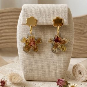 Boucles d'oreilles artisanales en forme de fleurs avec de véritables fleurs de glixia séchées figées dans la résine, montées sur des puces dorées en forme de fleur, présentées sur un support en lin.