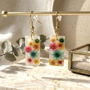Boucles d'oreilles rectangulaires artisanales en résine avec fleurs de glixia séchées colorées sur un support doré.