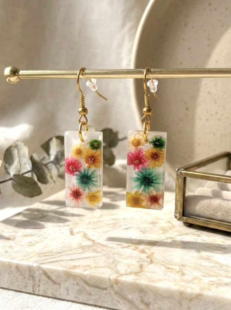Boucles d'oreilles rectangulaires artisanales en résine avec fleurs de glixia séchées colorées sur un support doré.