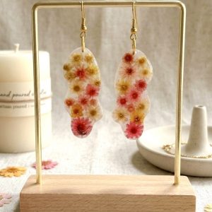 Boucles d'oreilles de la marque Clair de Maya en résine transparente de forme organique et irrégulière, renfermant de petites fleurs de glixia séchées aux tons jaune, orange et corail. Les boucles sont présentées sur un support doré avec en arrière-plan un tissu clair et une bougie.