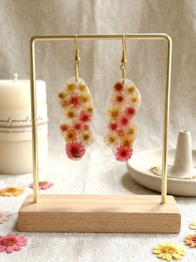 Boucles d'oreilles de la marque Clair de Maya en résine transparente de forme organique et irrégulière, renfermant de petites fleurs de glixia séchées aux tons jaune, orange et corail. Les boucles sont présentées sur un support doré avec en arrière-plan un tissu clair et une bougie.