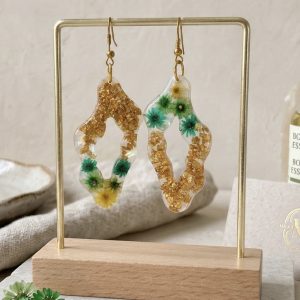 Une photographie montrant une paire de boucles d'oreilles artisanales uniques de la marque Clair de Maya, suspendues sur un support doré élégant avec une base en bois. Les boucles d'oreilles sont confectionnées en résine époxy transparente avec une forme organique asymétrique. À l'intérieur de la résine sont figées des fleurs de glixia séchées aux tons vert lagon et jaune, ainsi que des éclats de feuilles couleur or scintillants. Les fleurs et les dorures créent des motifs naturels et uniques. Les bijoux sont présentés dans un décor lumineux et esthétique sur un plateau en marbre, entourés de fleurs séchées dispersées et d'un brin de lavande, soulignant leur caractère naturel.