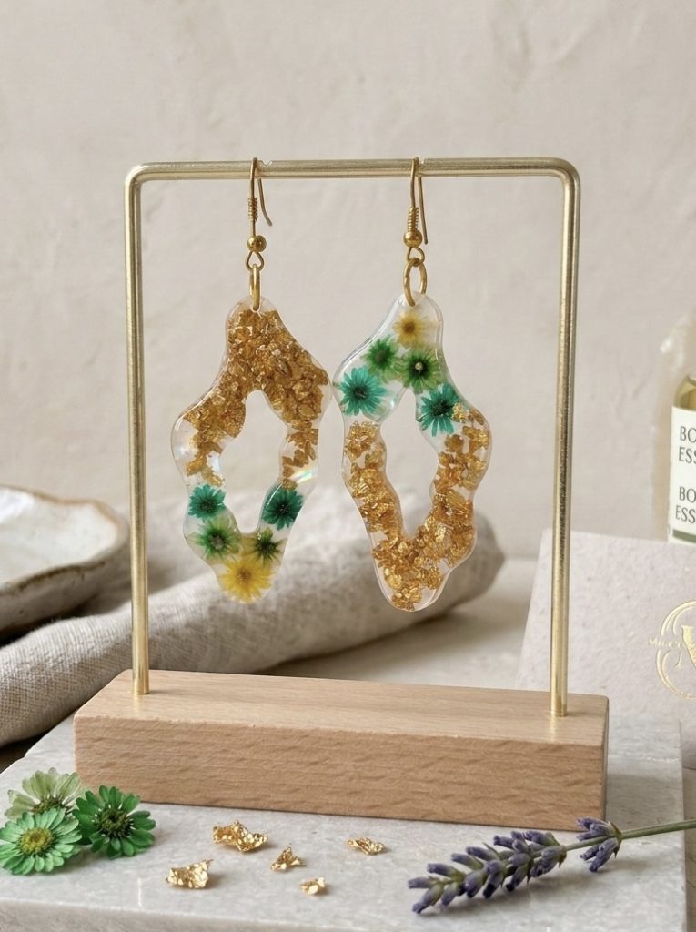 Une photographie montrant une paire de boucles d'oreilles artisanales uniques de la marque Clair de Maya, suspendues sur un support doré élégant avec une base en bois. Les boucles d'oreilles sont confectionnées en résine époxy transparente avec une forme organique asymétrique. À l'intérieur de la résine sont figées des fleurs de glixia séchées aux tons vert lagon et jaune, ainsi que des éclats de feuilles couleur or scintillants. Les fleurs et les dorures créent des motifs naturels et uniques. Les bijoux sont présentés dans un décor lumineux et esthétique sur un plateau en marbre, entourés de fleurs séchées dispersées et d'un brin de lavande, soulignant leur caractère naturel.