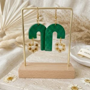 Boucles d'oreilles artisanales pendantes en forme d'arches vertes avec un pendentif en résine contenant de petites fleurs de glixia séchées, présentées sur un support doré.