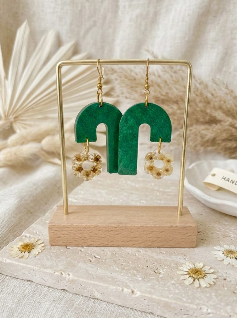 Boucles d'oreilles artisanales pendantes en forme d'arches vertes avec un pendentif en résine contenant de petites fleurs de glixia séchées, présentées sur un support doré.