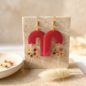 La photo présente une paire de boucles d'oreilles pendantes uniques et artisanales. Elles se composent de deux parties : une partie supérieure en résine d'un rose carmin intense en forme d'arches modernes asymétriques, et une partie inférieure suspendue en résine transparente renfermant de délicates fleurs de glixia séchées (tons jaunes, orangés et roses). Les attaches sont de couleur dorée. Les bijoux sont exposés sur un bloc rectangulaire en travertin beige, posé sur un plateau en marbre. En arrière-plan, on aperçoit une coupelle en céramique avec quelques fleurs séchées et un brin de pampa, le tout sous une lumière naturelle et douce.