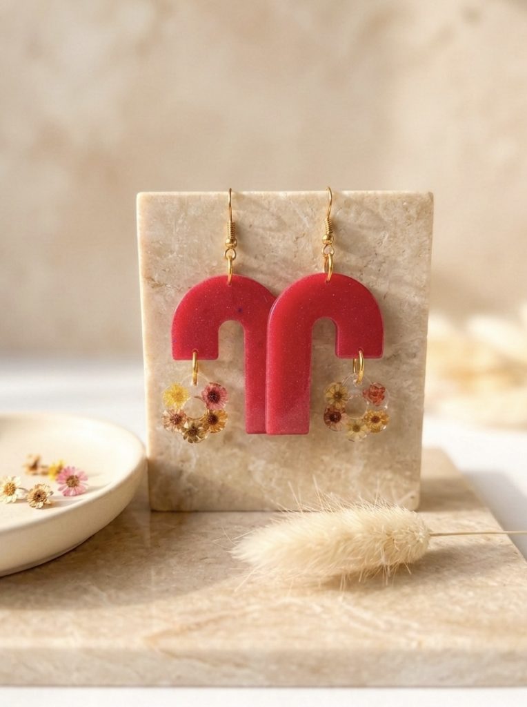 La photo présente une paire de boucles d'oreilles pendantes uniques et artisanales. Elles se composent de deux parties : une partie supérieure en résine d'un rose carmin intense en forme d'arches modernes asymétriques, et une partie inférieure suspendue en résine transparente renfermant de délicates fleurs de glixia séchées (tons jaunes, orangés et roses). Les attaches sont de couleur dorée. Les bijoux sont exposés sur un bloc rectangulaire en travertin beige, posé sur un plateau en marbre. En arrière-plan, on aperçoit une coupelle en céramique avec quelques fleurs séchées et un brin de pampa, le tout sous une lumière naturelle et douce.