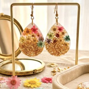 Boucles d'oreilles pendantes en forme de gouttes, réalisées en résine transparente. À l'intérieur sont figées de petites fleurs séchées multicolores (rose, jaune, bleu) et des éclats de feuilles d'or à la base. Les boucles sont présentées sur un support doré avec un miroir et des fleurs printanières en arrière-plan.