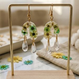 Photographie de produit d'une paire de boucles d'oreilles uniques de la marque Clair de Maya. Les boucles sont pendantes, en forme d'arches inversées en résine transparente, dans lesquelles sont incrustées de petites fleurs de glixia séchées à la main dans des tons vert turquoise et jaune doré. Sous chaque arche pendent deux gouttes cristallines. Le bijou est suspendu sur un support métallique de couleur dorée, posé sur un comptoir en marbre. En arrière-plan, on aperçoit une serviette en lin, des fleurs séchées éparpillées et des pompons en laine blanche.