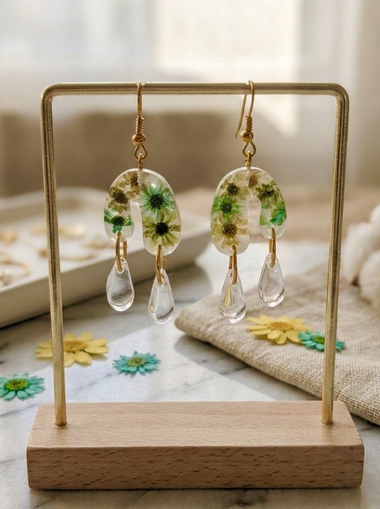 Photographie de produit d'une paire de boucles d'oreilles uniques de la marque Clair de Maya. Les boucles sont pendantes, en forme d'arches inversées en résine transparente, dans lesquelles sont incrustées de petites fleurs de glixia séchées à la main dans des tons vert turquoise et jaune doré. Sous chaque arche pendent deux gouttes cristallines. Le bijou est suspendu sur un support métallique de couleur dorée, posé sur un comptoir en marbre. En arrière-plan, on aperçoit une serviette en lin, des fleurs séchées éparpillées et des pompons en laine blanche.