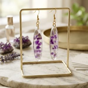 Gros plan sur une paire de boucles d'oreilles pendantes de forme ovale, suspendues sur un support doré minimaliste. Les bijoux sont créés en résine transparente incluant des éclats d'améthyste naturelle d'un violet profond. L'ensemble est mis en scène sur un plateau en marbre blanc, entouré de géodes d'améthyste brute, d'un brin de lavande séchée et d'un flacon de parfum, le tout baigné dans une lumière naturelle douce.