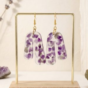 Cette photographie présente une paire de boucles d'oreilles artisanales en forme d'arches asymétriques, créées par la marque Clair de Maya. Chaque boucle est une pièce unique en résine transparente, à l'intérieur de laquelle sont encapsulés de multiples fragments d'améthyste naturelle violette, aux formes brutes et facettées. La disposition irrégulière des pierres souligne leur caractère organique et exclusif. Les boucles sont suspendues à de délicats crochets de couleur or poli. Elles sont présentées sur un support métallique doré minimaliste avec un socle en bois clair. En arrière-plan, des brins de lavande séchée et des cristaux d'améthyste brute reposent sur une surface texturée blanche, créant une ambiance naturelle, douce et harmonieuse.