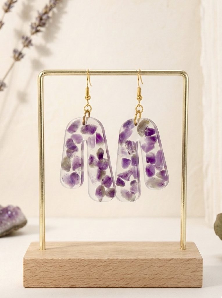 Cette photographie présente une paire de boucles d'oreilles artisanales en forme d'arches asymétriques, créées par la marque Clair de Maya. Chaque boucle est une pièce unique en résine transparente, à l'intérieur de laquelle sont encapsulés de multiples fragments d'améthyste naturelle violette, aux formes brutes et facettées. La disposition irrégulière des pierres souligne leur caractère organique et exclusif. Les boucles sont suspendues à de délicats crochets de couleur or poli. Elles sont présentées sur un support métallique doré minimaliste avec un socle en bois clair. En arrière-plan, des brins de lavande séchée et des cristaux d'améthyste brute reposent sur une surface texturée blanche, créant une ambiance naturelle, douce et harmonieuse.
