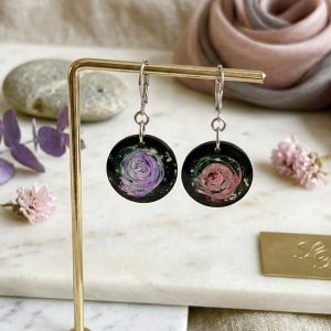 La photo présente une paire de boucles d'oreilles pendantes circulaires de la collection artisanale Galaxy, disposées sur un support doré. Chaque boucle présente un fond noir avec un motif tourbillonnant incrusté dans la résine : l'une dans les tons violets, l'autre dans un rose poudré avec des éclats de poussière d'étoiles. Le fond de l'image est clair, en marbre, avec des éléments décoratifs tels que des fleurs séchées et un tissu soyeux, soulignant le caractère artisanal du bijou.
