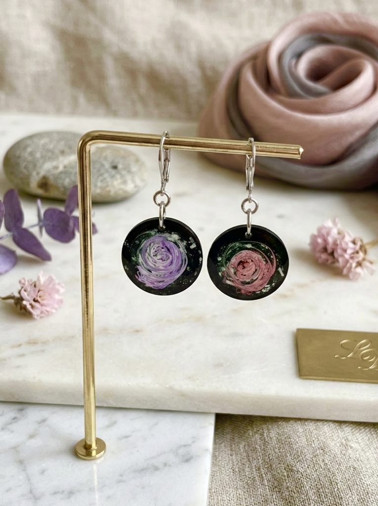 La photo présente une paire de boucles d'oreilles pendantes circulaires de la collection artisanale Galaxy, disposées sur un support doré. Chaque boucle présente un fond noir avec un motif tourbillonnant incrusté dans la résine : l'une dans les tons violets, l'autre dans un rose poudré avec des éclats de poussière d'étoiles. Le fond de l'image est clair, en marbre, avec des éléments décoratifs tels que des fleurs séchées et un tissu soyeux, soulignant le caractère artisanal du bijou.