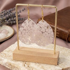 Boucles d'oreilles artisanales en forme de grandes gouttes, remplies de petits éclats de quartz rose naturel sertis dans une résine transparente. Les boucles sont suspendues sur un support doré, avec de la lavande et un carnet en cuir en arrière-plan.
