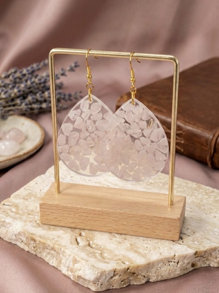 Boucles d'oreilles artisanales en forme de grandes gouttes, remplies de petits éclats de quartz rose naturel sertis dans une résine transparente. Les boucles sont suspendues sur un support doré, avec de la lavande et un carnet en cuir en arrière-plan.