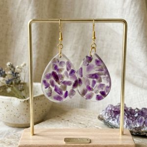 Boucles d'oreilles en forme de gouttes en résine transparente avec des éclats d'améthyste naturelle violette à l'intérieur, suspendues sur un support doré devant un tissu clair et de la lavande.