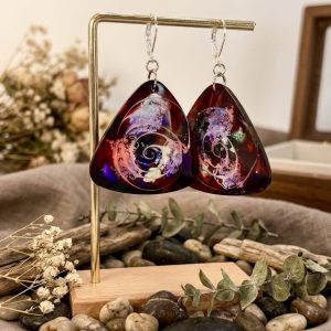 Une paire de boucles d'oreilles triangulaires en résine époxy de la collection Galaxy, suspendues sur un support doré. L'intérieur de la résine révèle des volutes tourbillonnantes de violet, de rose et de blanc sur un fond bordeaux profond, évoquant une nébuleuse cosmique. Des pierres naturelles et des plantes séchées sont disposées à la base de la composition.