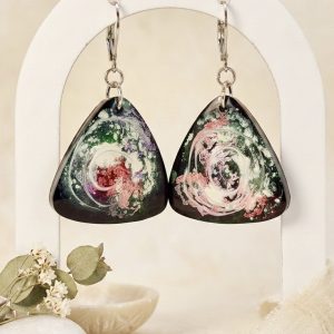 Boucles d'oreilles uniques en résine époxy en forme de triangles, issues de la collection Galaxy. L'intérieur de la résine sombre révèle des nébuleuses tourbillonnantes aux tons vert, blanc et rose. Les boucles sont montées sur des dormeuses argentées en acier chirurgical, présentées sur un support blanc avec des éléments naturels tels que des fleurs séchées et des pierres.