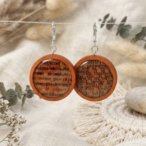 Boucles d'oreilles pendantes rondes en bois clair, incrustées de fragments de pages de vieux livres sous une résine transparente, présentées sur un fond de tissus beiges et de fleurs séchées.