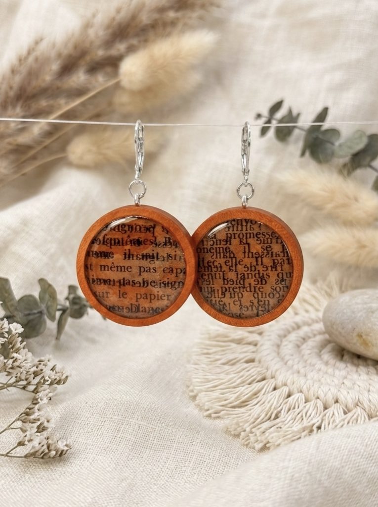 Boucles d'oreilles pendantes rondes en bois clair, incrustées de fragments de pages de vieux livres sous une résine transparente, présentées sur un fond de tissus beiges et de fleurs séchées.